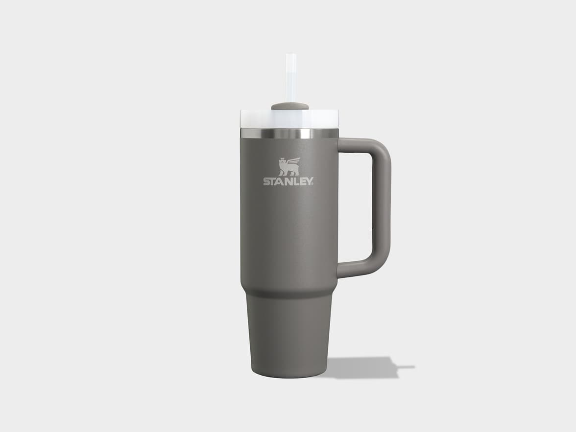 A tall, gray Stanley brand tumbler with a handle and a clear straw, standing upright on a light gray surface. The Stanley logo is visible on the front.