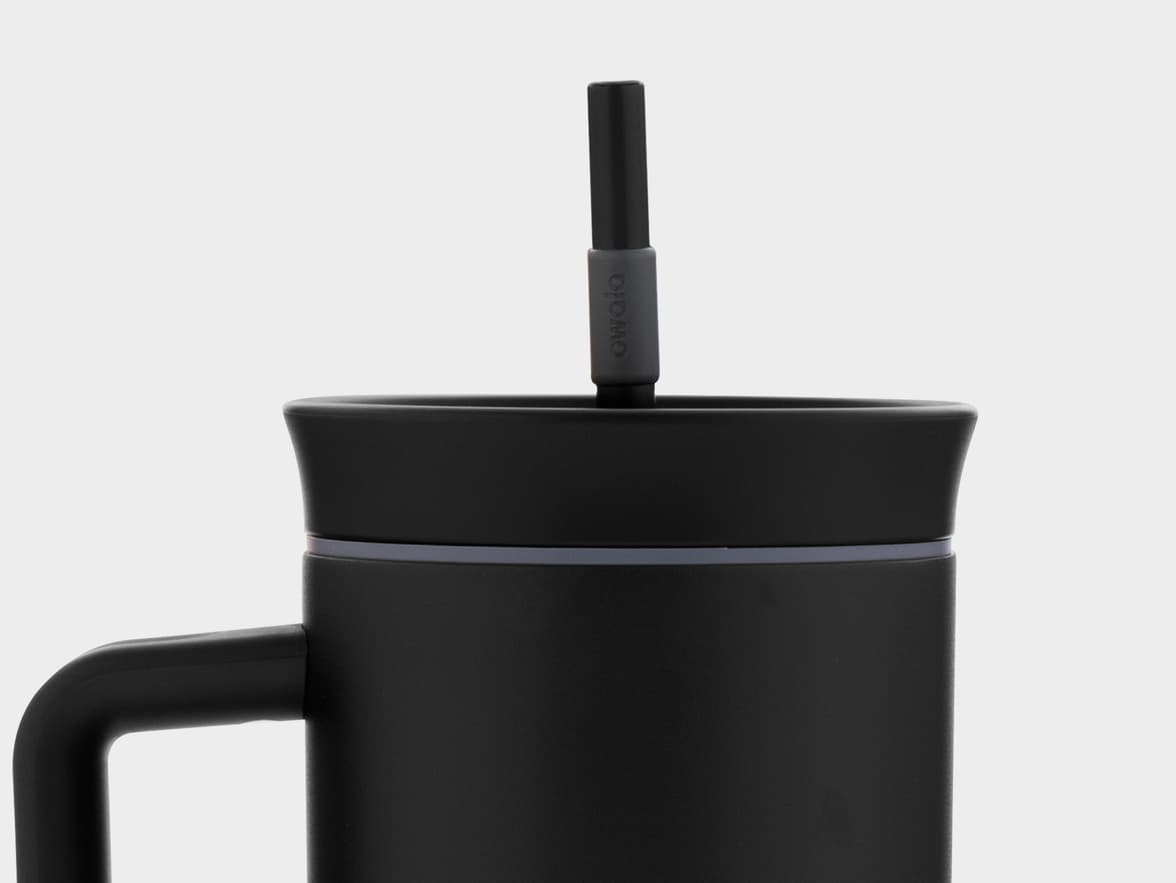 A close-up shot of a black Owala travel mug with a handle, lid, and a straw extending upwards from the lid, against a plain light grey background.