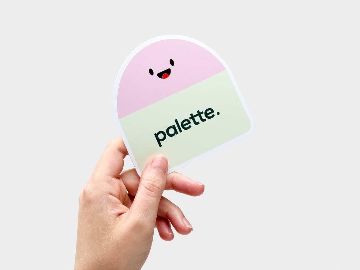 A hand holding a customized coated sticker of a rectangle logo with a half circle face on top.