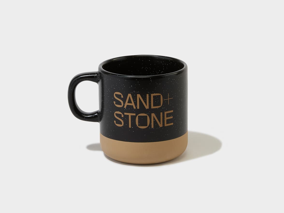 A black speckled mug with a tan bottom, branded with 'SAND+STONE' in gold lettering.