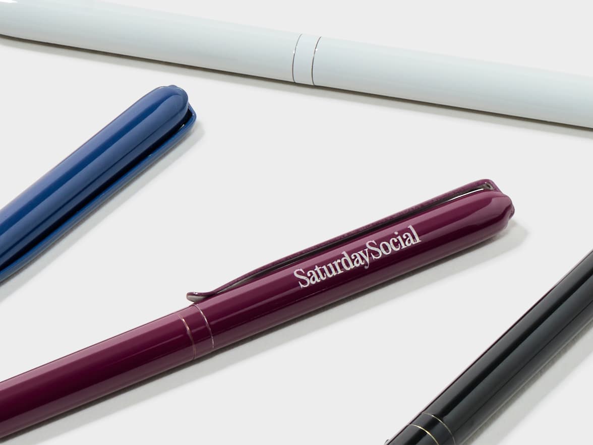 Four pens in white, blue, mulberry, and black on a white background. The mulberry pen features "SaturdaySocial" branding.