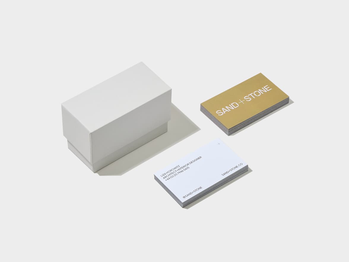 White business cards with a gold-colored edge, one with "SAND + STONE" in white on a gold background, and another with "LISA HOROWITZ, ARCHITECT + INTERIOR DESIGNER +44 (0) 20 7946 0615" and "@SAND+STONE" on a white background, next to a white box.