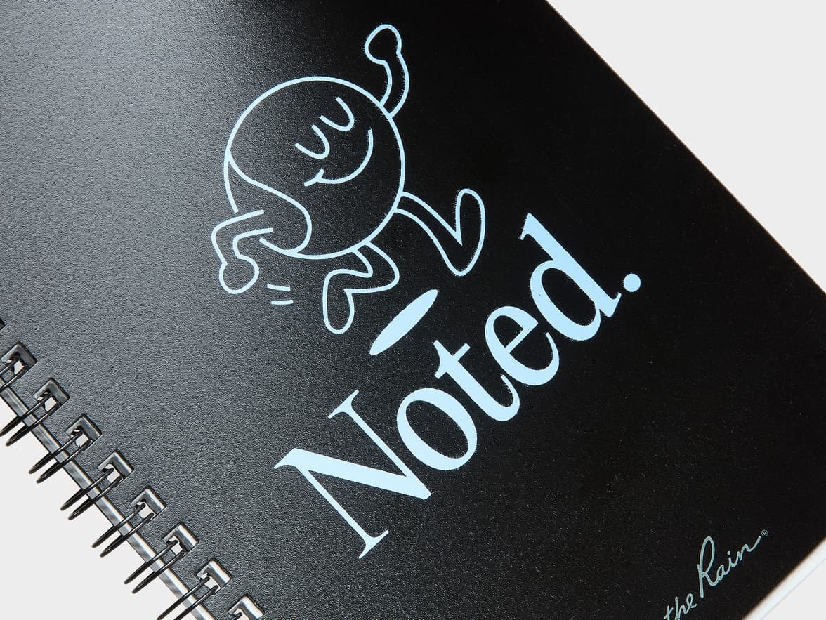 A black spiral-bound notebook with a whimsical line-drawn character and the branding "Noted. the Rain" on its textured cover.