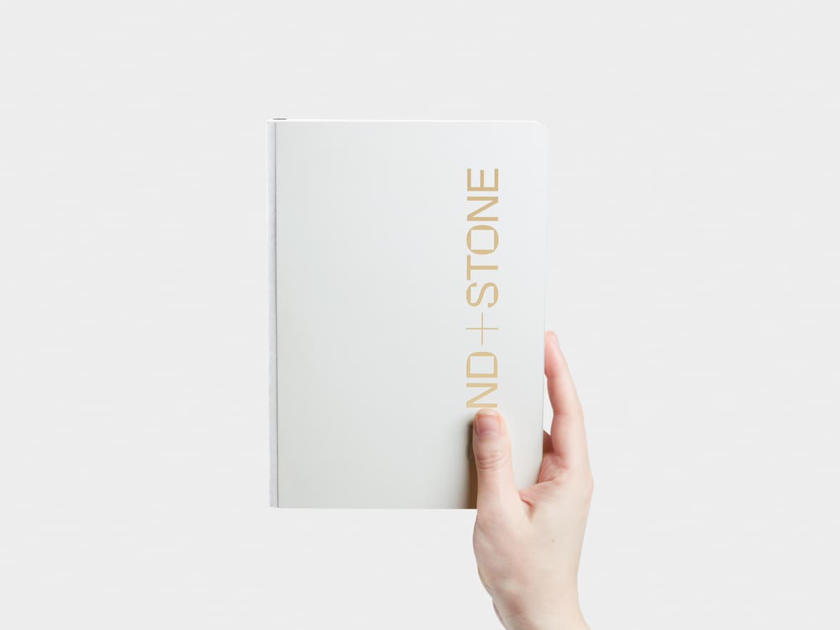 A hand holding a cloudy grey softcover notebook with gold foil customisation.