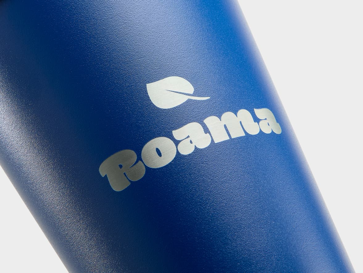 Close-up view of a matte navy blue insulated water bottle, angled slightly. The bottle has a subtle textured finish. Towards the top, a silver leaf logo is visible, followed by the word "Roama" in a rounded, silver font with a brushed metal appearance. The background is a soft, light grey.