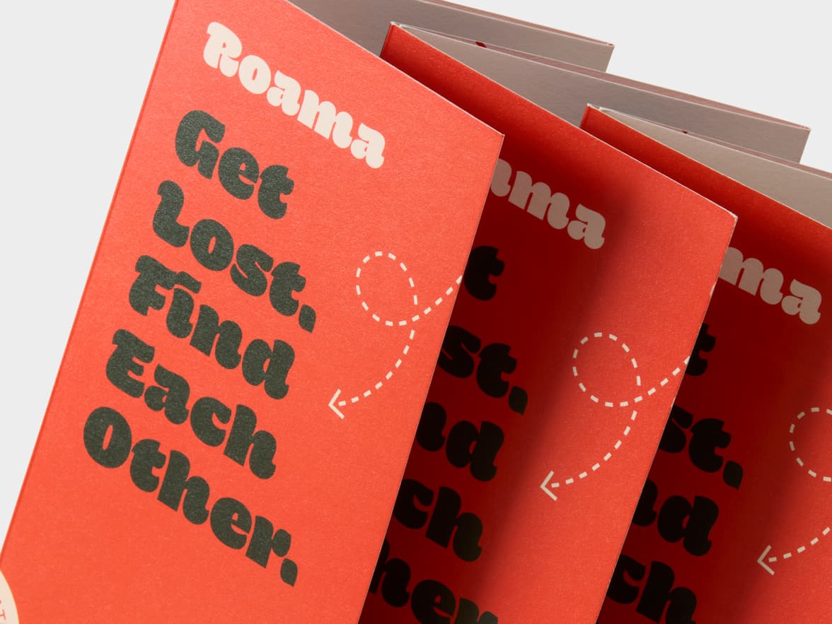 Fanned red booklets display "Roama" and "Get Lost. Find Each Other." with a dashed arrow graphic.