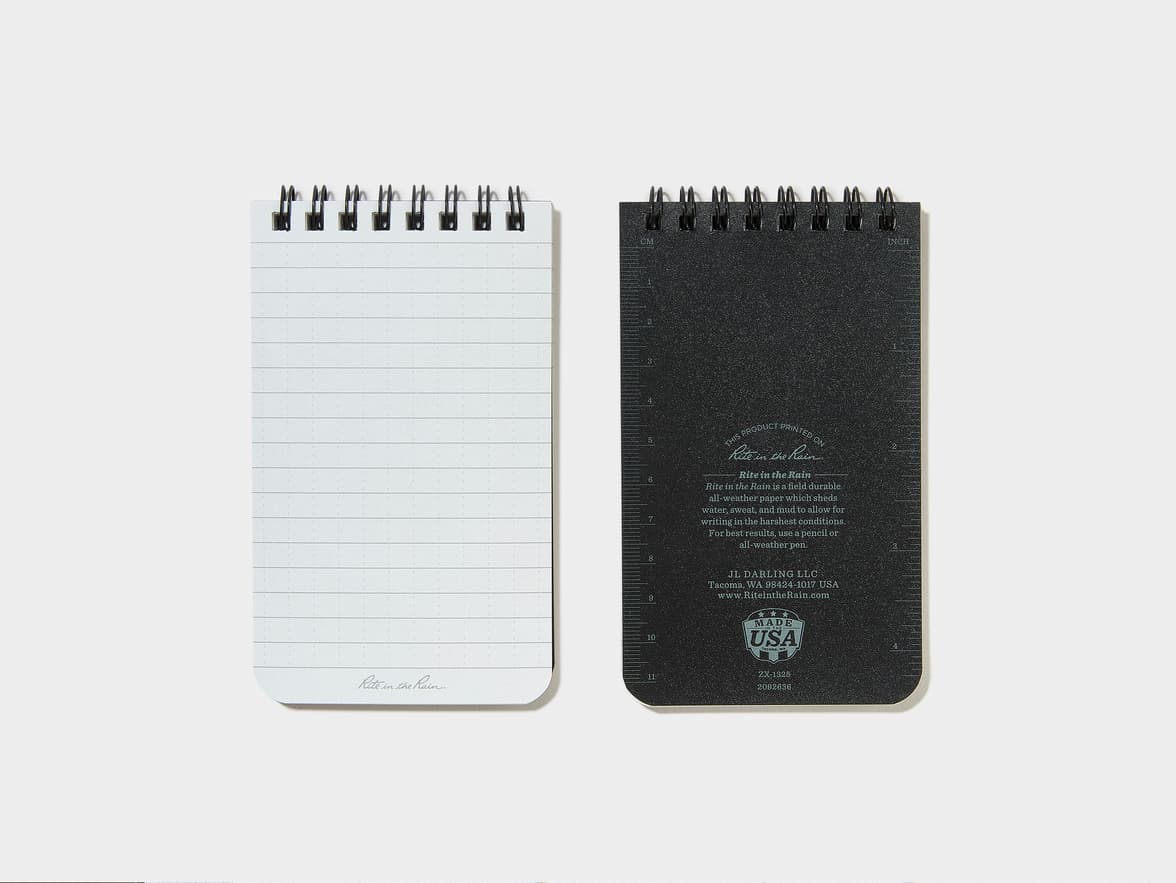 A Rite in the Rain spiral-bound notepad, showing its white lined paper front and black textured back cover with branding text including 'Made in the USA'.