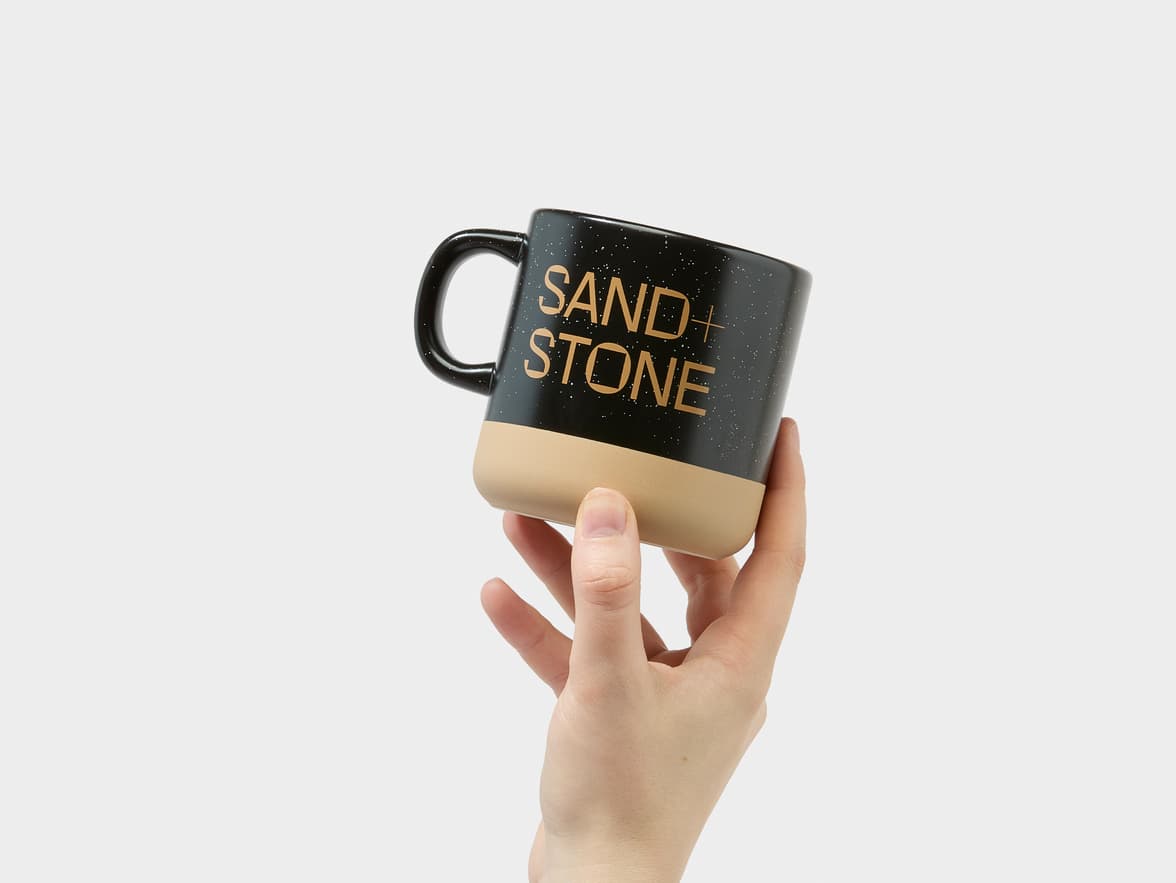 A hand holds a black and beige mug with "SAND + STONE" written in gold letters.