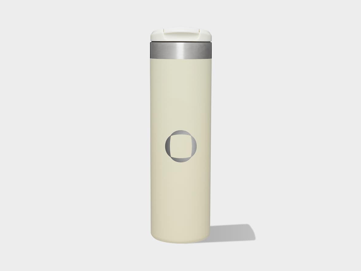 A cream-colored insulated travel mug with a white lid, a silver band, and a silver circular logo featuring a square in its center.