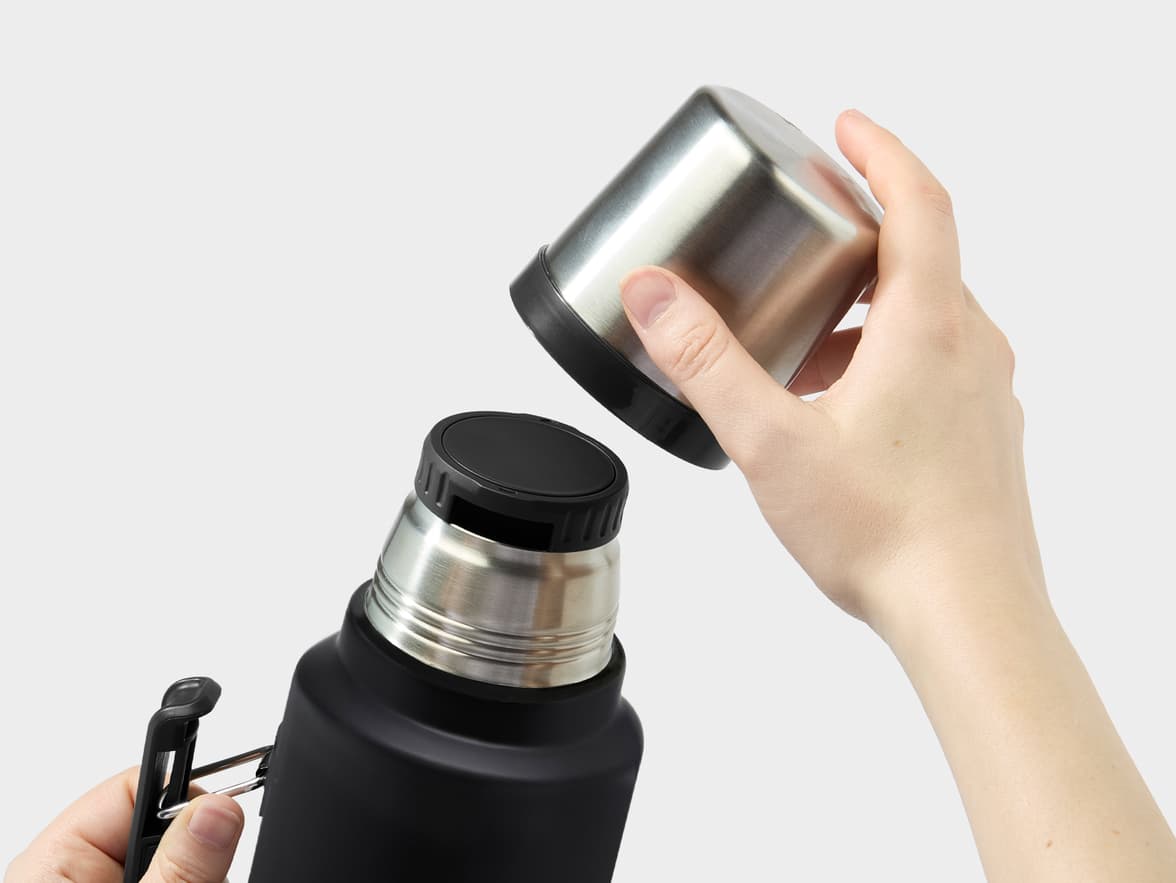 A person's hands opening a black thermos bottle with a stainless steel cup.