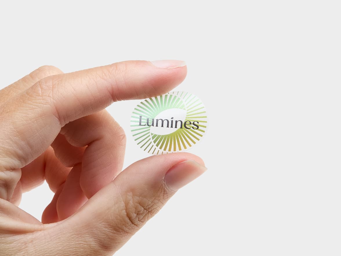 A hand holding a customized round clear sticker with a light green spiral circle and the word 'Lumines' printed on it.