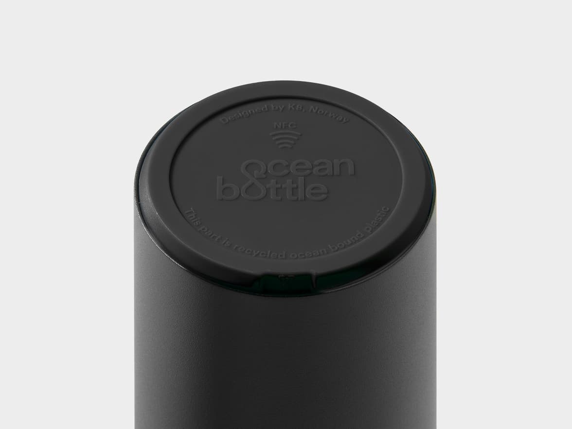 A close-up of the black lid of an 'ocean bottle' water bottle. The lid is embossed with "Designed by KB, Norway", an NFC logo, the "ocean bottle" logo, and "This part is recycled ocean bound plastic".