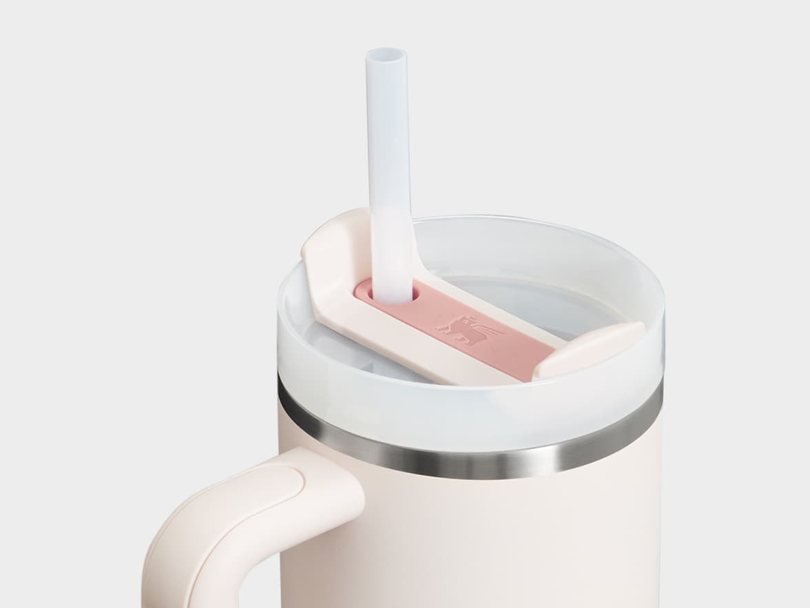A close-up view of the top part of a cream-colored Stanley Quencher H2.0 FlowState Tumbler, showing the clear lid, a translucent white straw, and a pink rotating cover on the lid with a detailed embossed Stanley bear logo. A light-colored handle is visible on the left side of the tumbler.