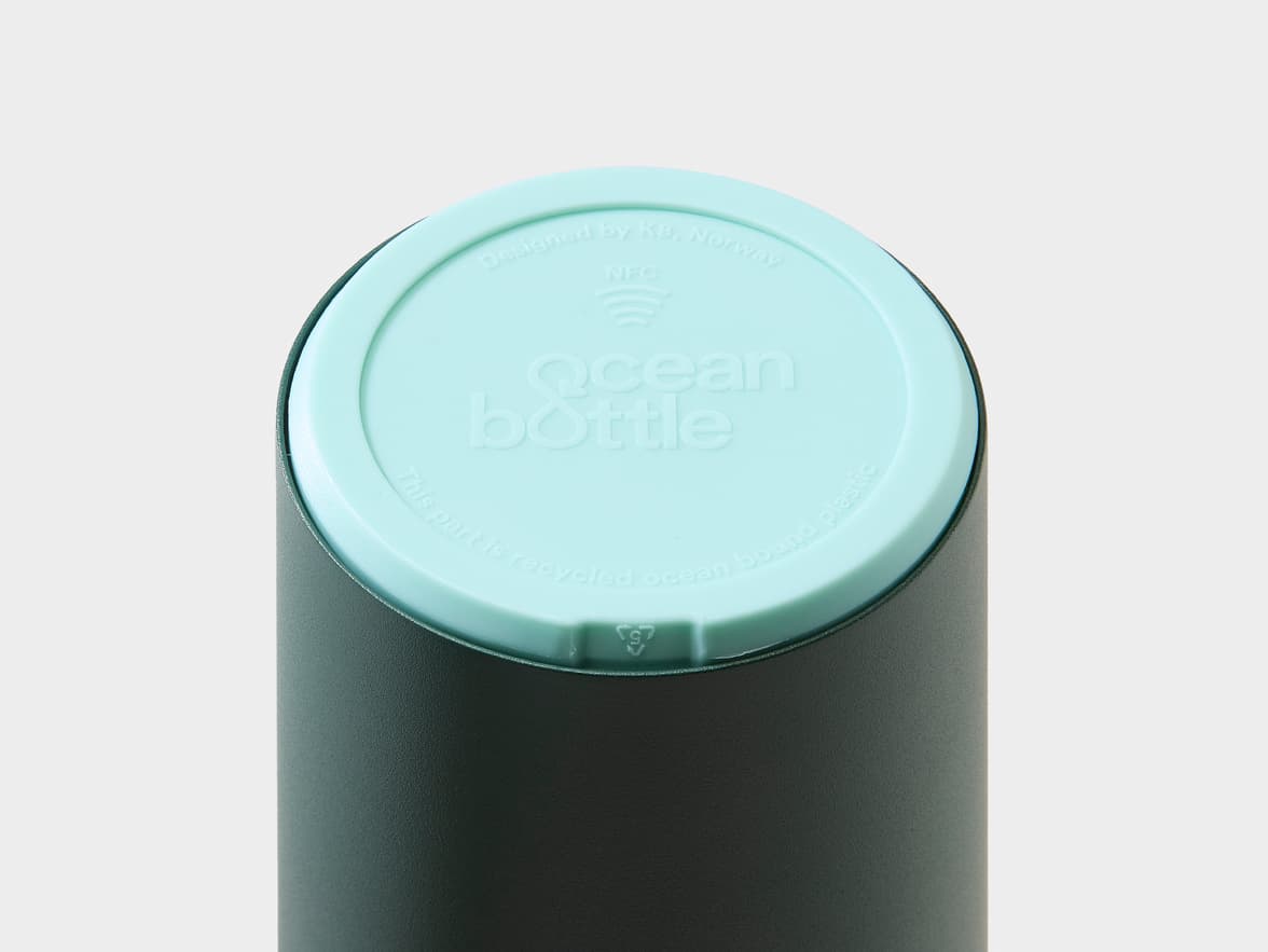 A dark green Ocean Bottle with a light blue cap. The cap displays the branding 'ocean bottle', along with text 'Designed by KB, Norway', 'NFC', and 'This part is recycled ocean bound plastic'.