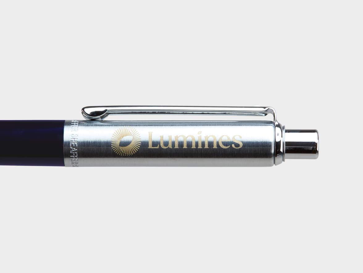 Close-up of a dark blue Sheaffer pen with a brushed metal cap. The cap features a golden sunburst logo and "Lumines" engraved horizontally, and "SHEAFFER" engraved vertically.