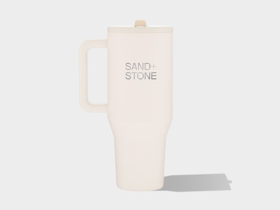 A cream-colored travel mug with a handle and a lid, featuring the text 'SAND+STONE' printed vertically in metallic gray, stands upright on a plain light gray surface, casting a shadow to its right.