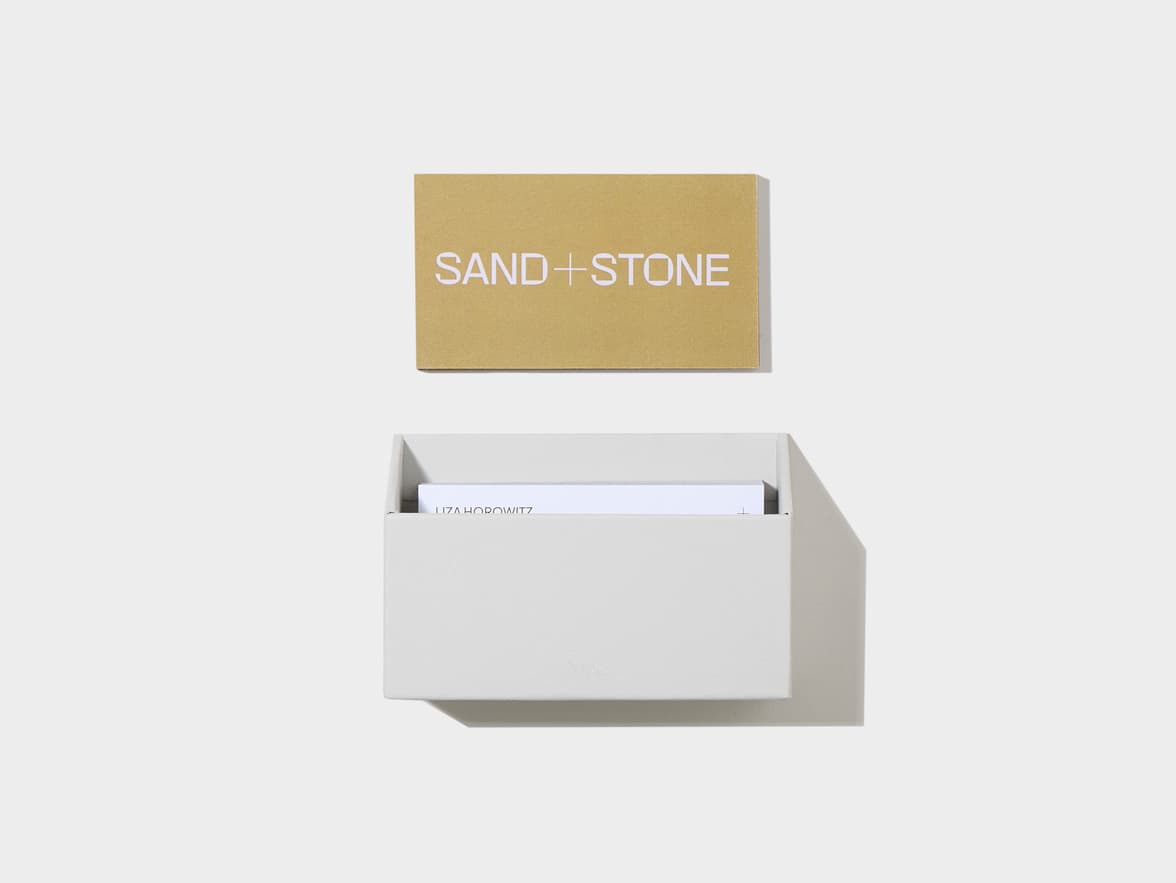 A gold card with "SAND + STONE" placed above a light grey card holder containing white cards displaying "LIZA HOROWITZ" and a droplet logo.