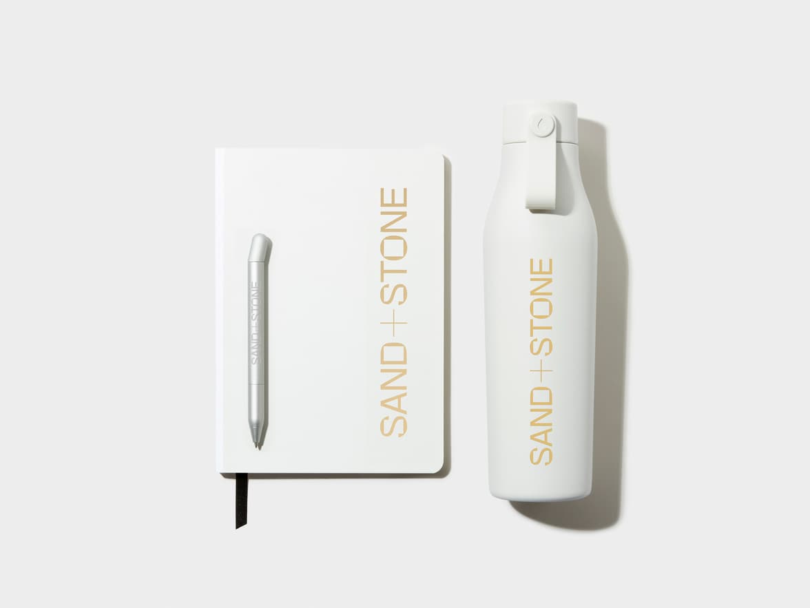 A cloudy grey softcover notebook with gold foil customisation, alongside a cloudy grey custom pen and water bottle.
