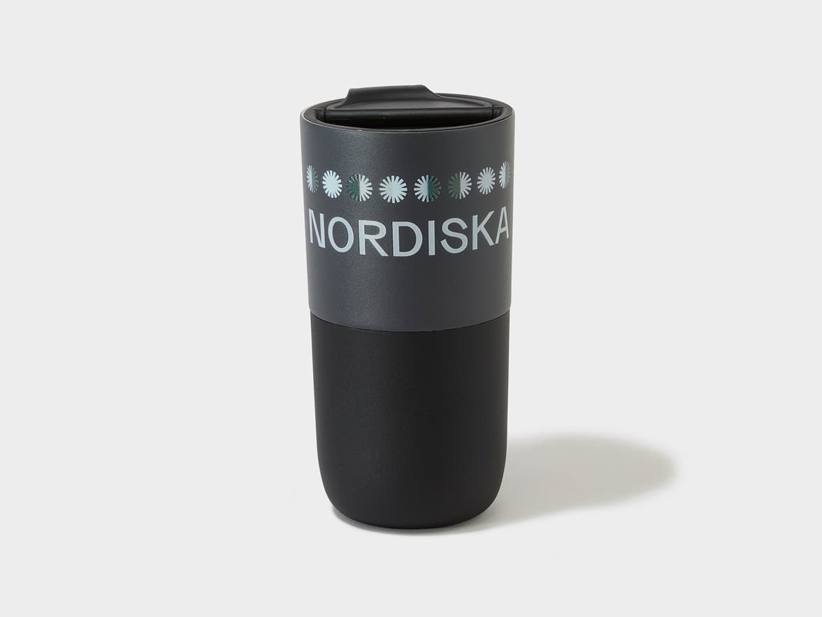 A dark gray and black tumbler with "NORDISKA" written in light gray letters and a band of light gray and green stylized snowflakes/flowers above the text.