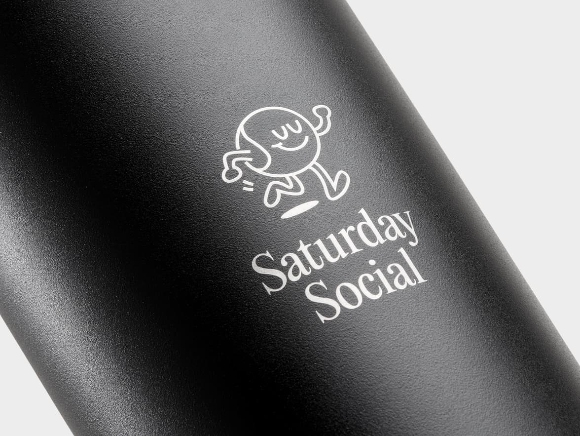 A close-up shot of a black, textured cylindrical object, likely a tumbler or bottle, featuring a white graphic and text. The graphic is a whimsical outline drawing of a smiling, dancing character with small arms and legs. Below it, the words "Saturday Social" are printed in a white serif font. The object has a matte, granular finish and is lit from the right, creating a gradient from lighter gray to darker black.