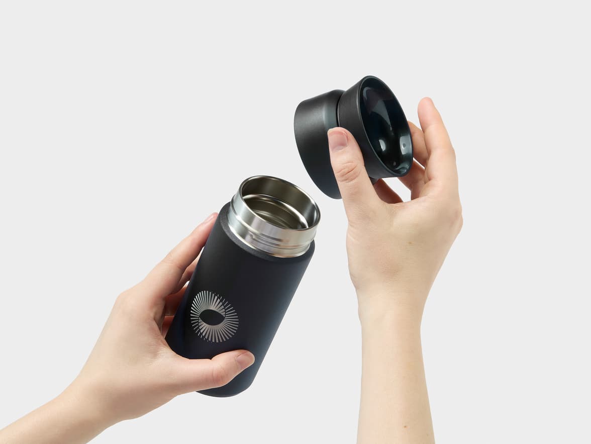 A person's hands hold an open black thermal bottle with a silver, stylized circular logo featuring radiating lines.