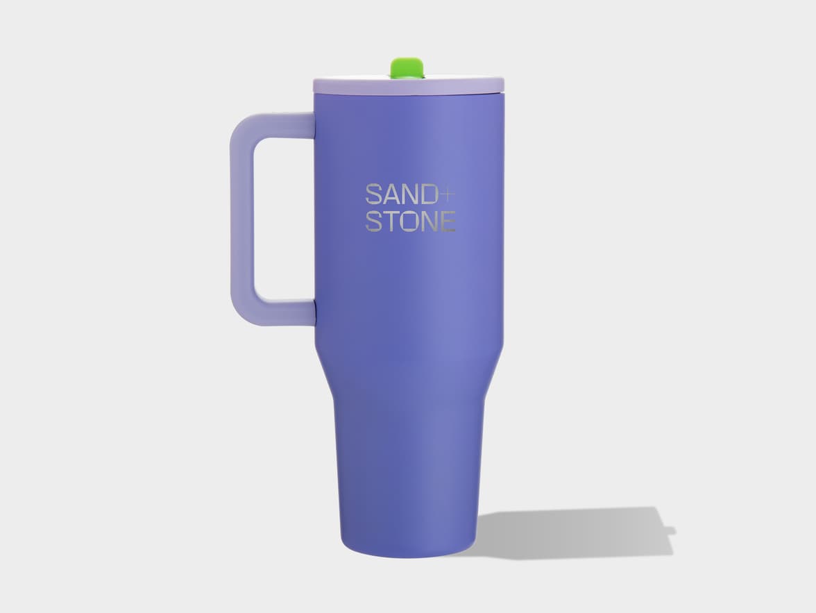 A matte purple travel mug with a matching handle, a light purple lid, and a green sip tab. The brand "SAND + STONE" is printed vertically on the front. The mug is on a plain light background.