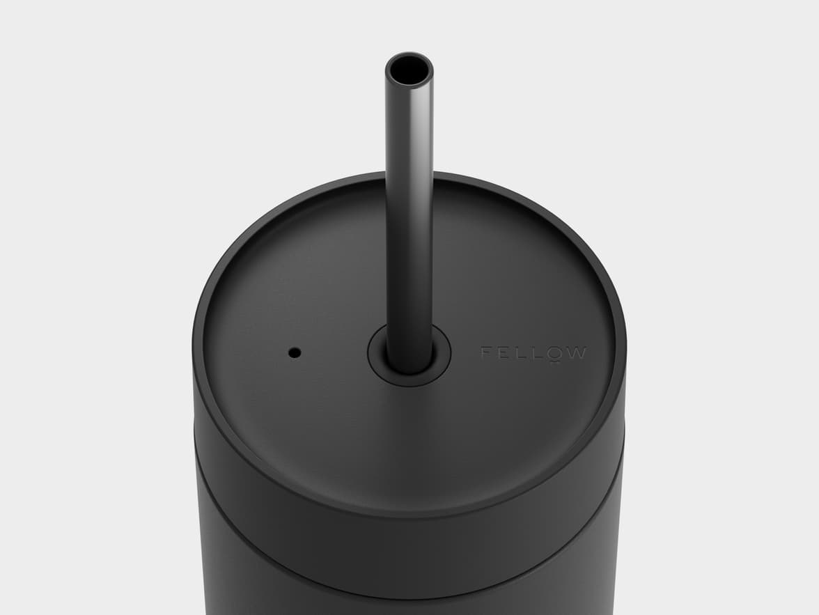 A top-down view of a matte black tumbler with a black straw. The straw is centered on the black lid, which also has a small circular vent hole to the left and the word "FELLOW" embossed to the right. The top portion of the tumbler's cylindrical body is visible below the lid, all set against a light gray background.