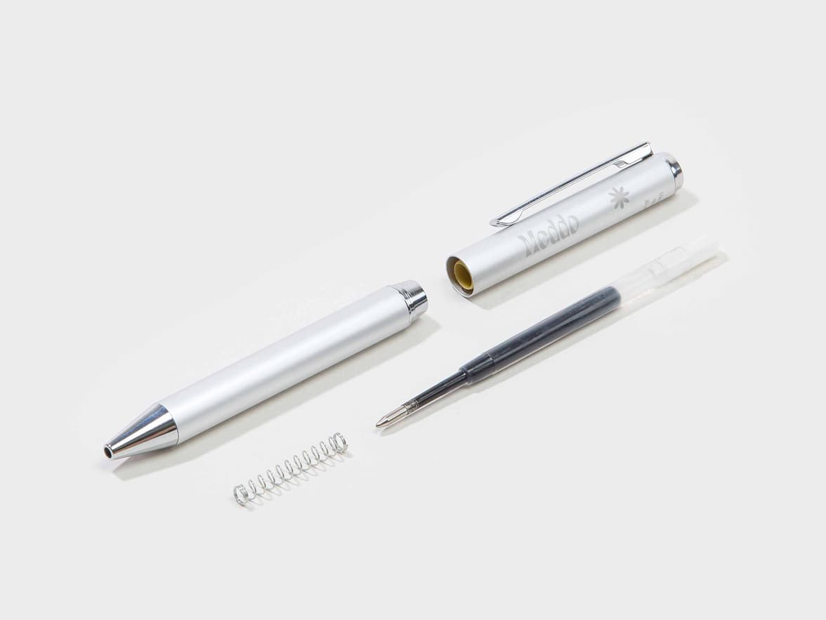 A silver Meddo brand pen disassembled, showing the main barrel with the "Meddo" logo and an asterisk, the tip, a spring, and a black ink refill.
