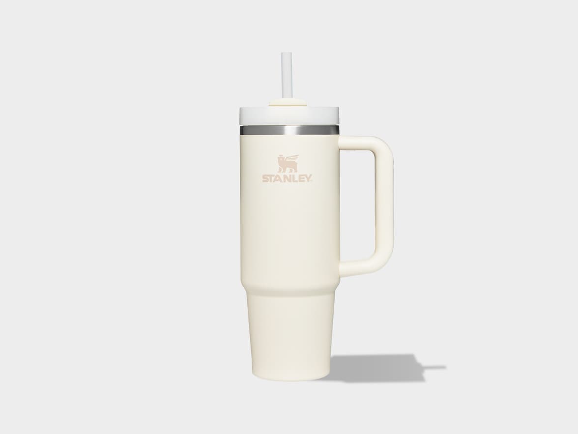 A cream-colored Stanley Quencher H2.0 FlowState tumbler with a handle and clear straw is centered on a light gray background, casting a subtle shadow to its right. The Stanley logo is visible on the front of the tumbler.