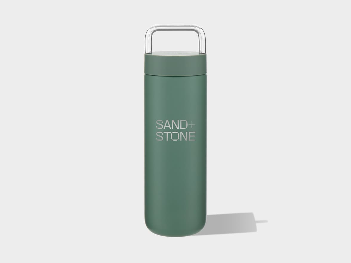 A muted green cylindrical bottle with a clear, U-shaped handle on top, features the silver text "SAND + STONE" vertically aligned on its side, standing on a light grey surface with a soft shadow to its right.