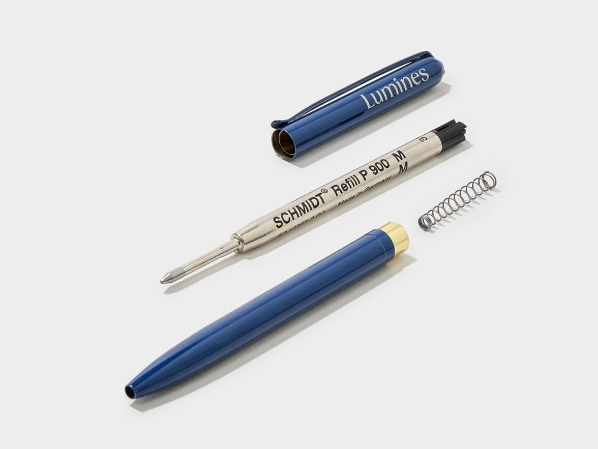 A disassembled blue Lumines ballpoint pen, showing its cap, barrel with a gold tip, a Schmidt P 900 M refill, and a small spring on a white background.