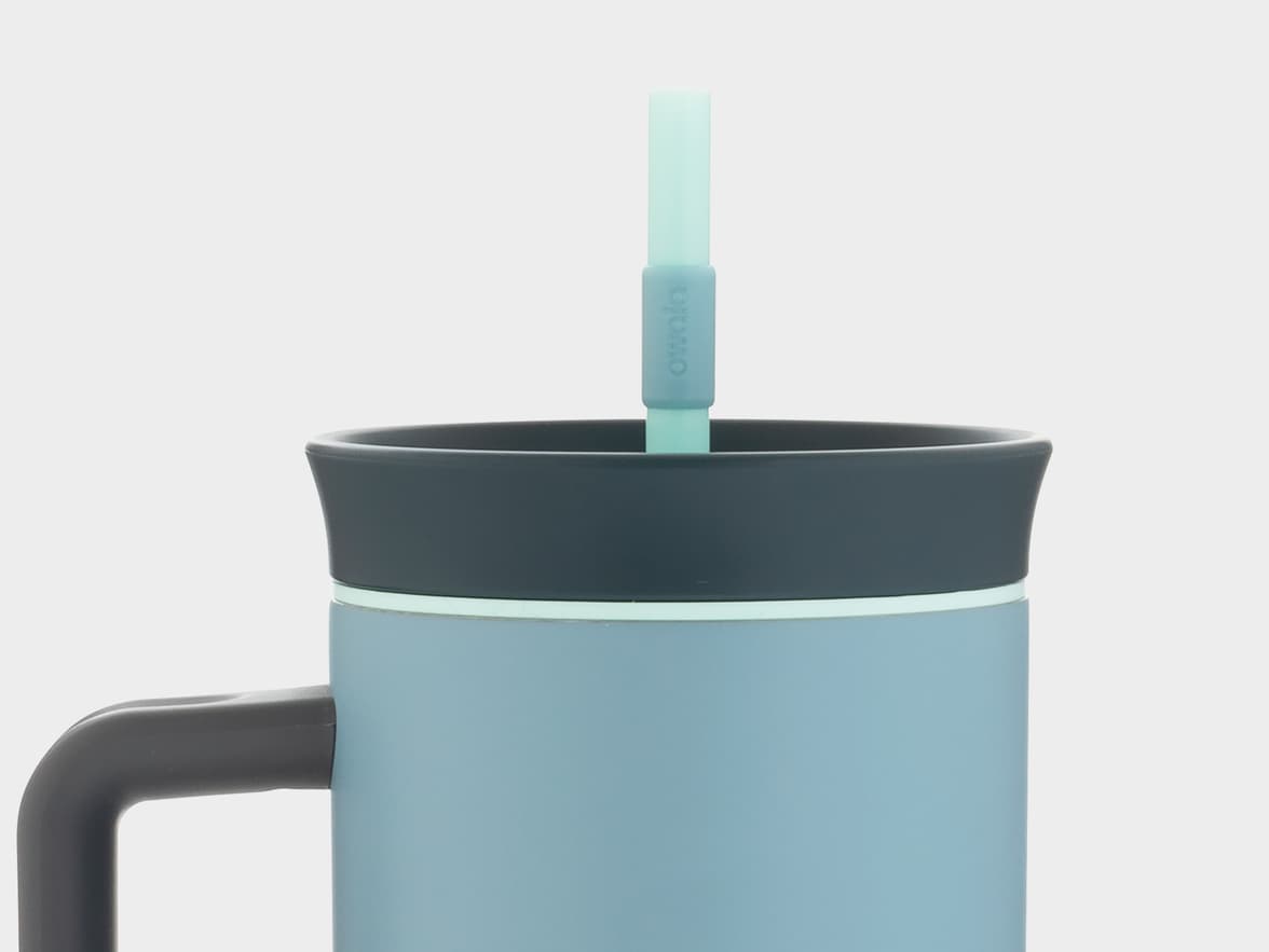 A close-up view of a light blue travel mug with a dark grey lid and handle, featuring a light blue straw with "owala" branding.