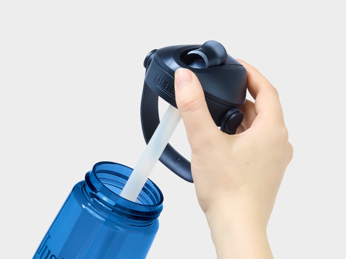A hand holds the dark blue CamelBak lid over a blue bottle, with a straw extending into the bottle.