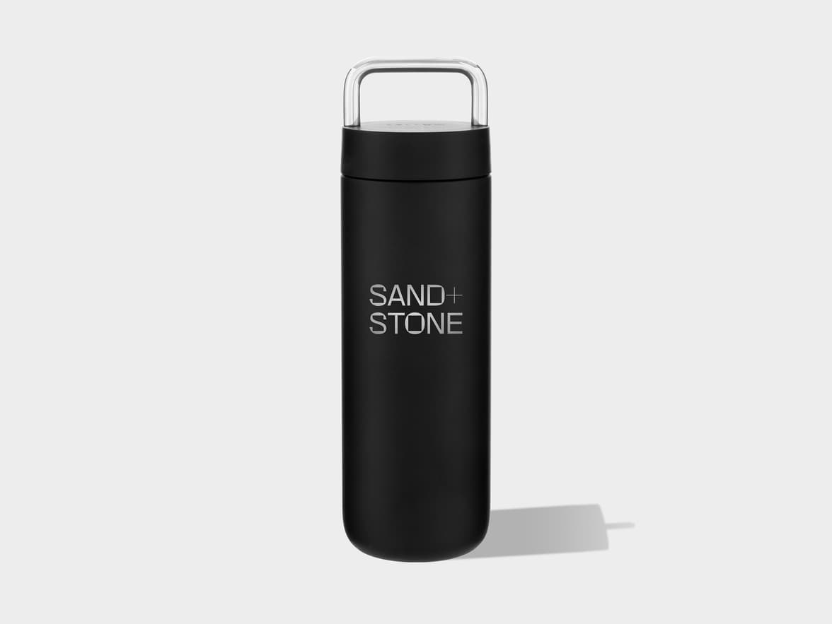Black travel mug with a clear, curved handle on top, standing upright on a light grey background, casting a soft shadow to the right. The words "SAND+STONE" are printed in light grey text on the side of the mug.