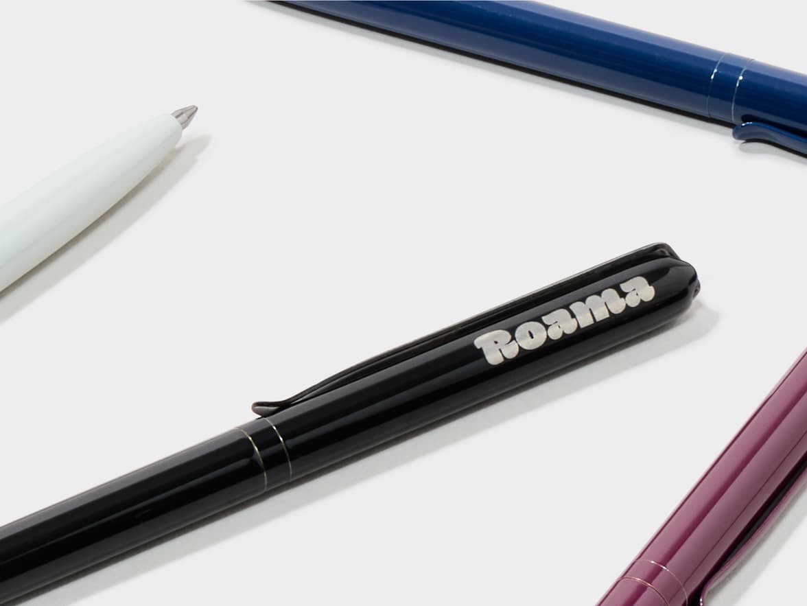 A black pen with "Roama" branding, a white pen, a blue pen, and a purple pen arranged on a white surface.