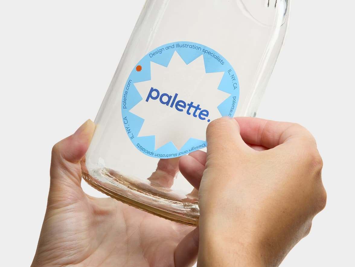 A hand placing a customized round clear sticker onto a glass bottle.