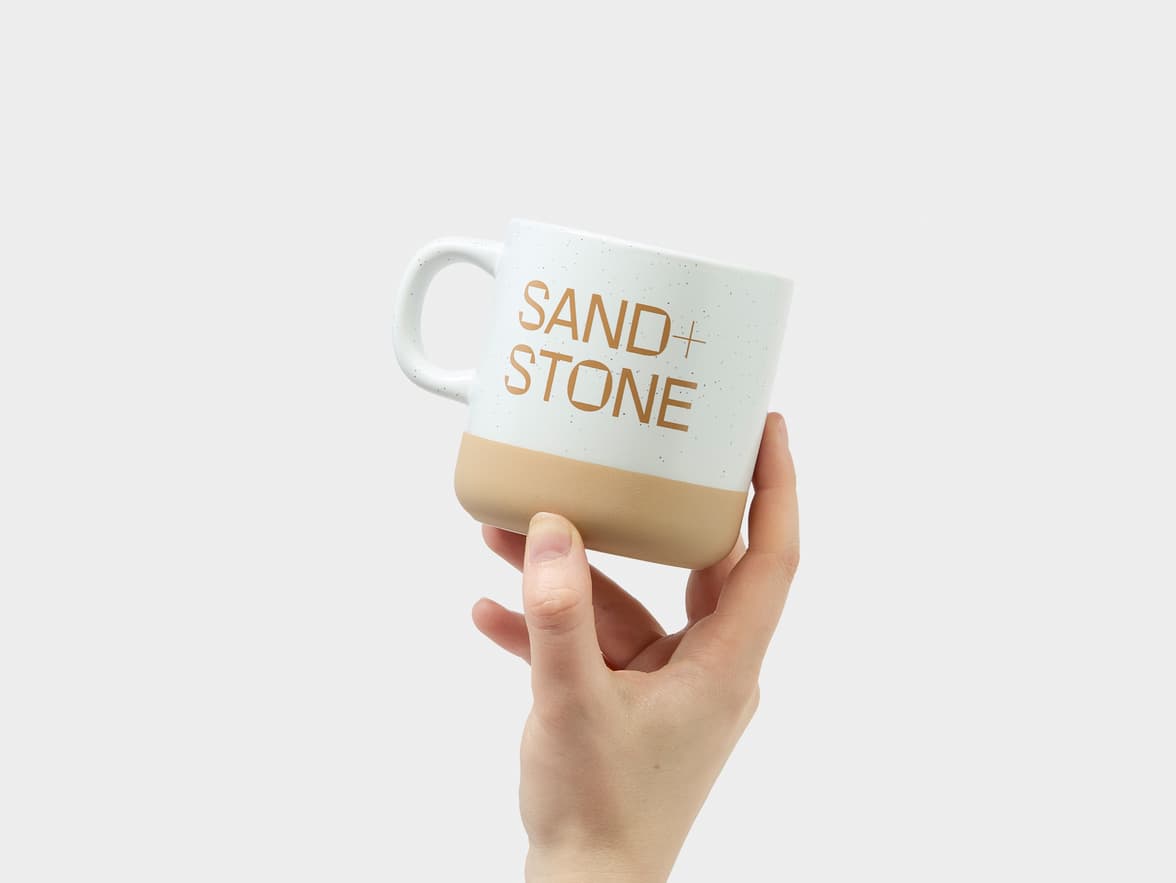 A hand holds a white and beige mug with the text "SAND + STONE" printed on it.