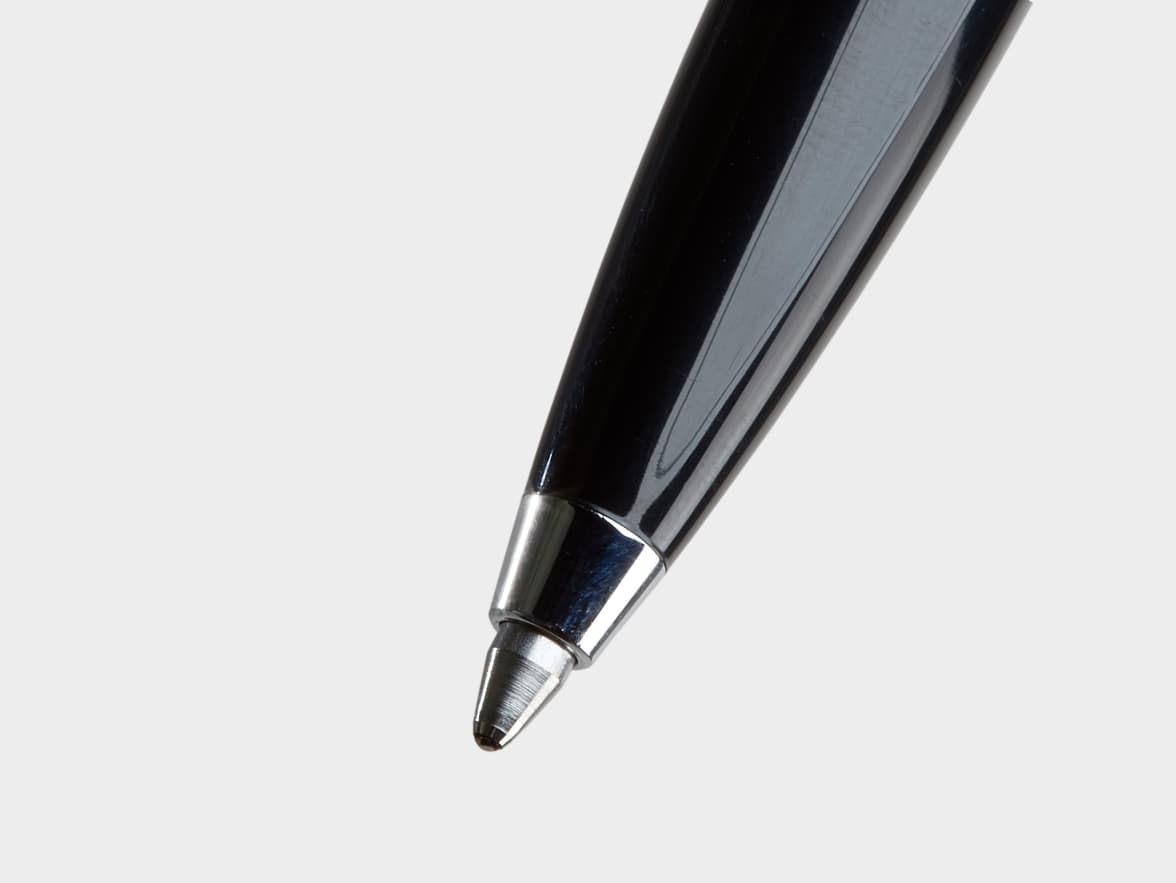 A close-up of a shiny black ballpoint pen with a silver tip, angled downwards on a light grey background.