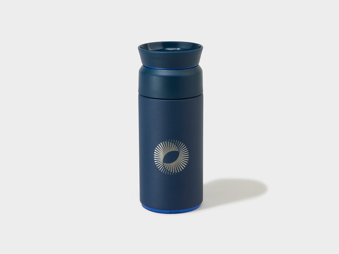 Navy blue travel mug with a silver circular logo featuring radiating lines, and a bright blue base.