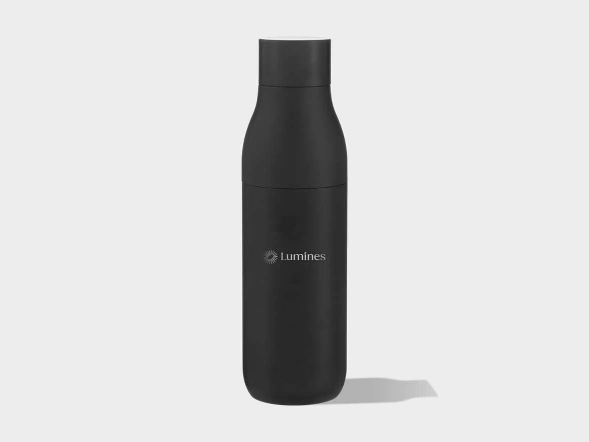 A tall, black, matte bottle with a white cap stands upright on a light grey surface, casting a subtle shadow. The bottle has a slight curve in the middle and features a light grey "Lumines" logo with a sunburst icon on its side.