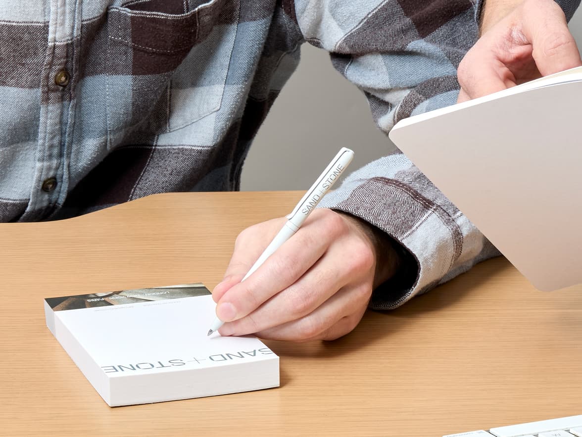Person writing on a SAND + STONE notepad with a SAND + STONE pen.
