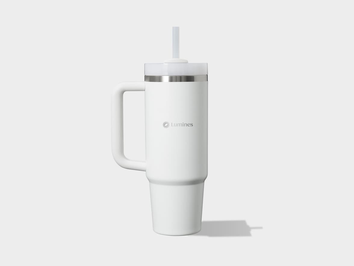 A tall, white, insulated tumbler with a handle on the left side and a clear straw protruding from the lid. A silver band is visible just below the lid, and the word "Lumines" with a sun-like logo is de-bossed on the front. 
