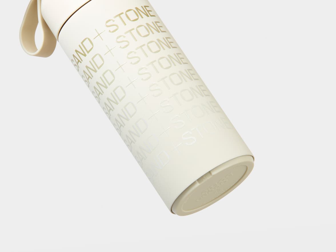 An off-white bottle with "SAND + STONE" branding and a loop handle.