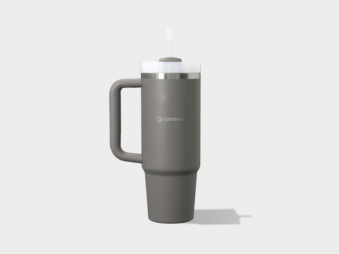 A gray tumbler with a handle stands on a light gray surface, casting a shadow to its right. The tumbler has a white lid and a clear straw protruding from the top. A "Lumines" logo is visible on the front of the tumbler.