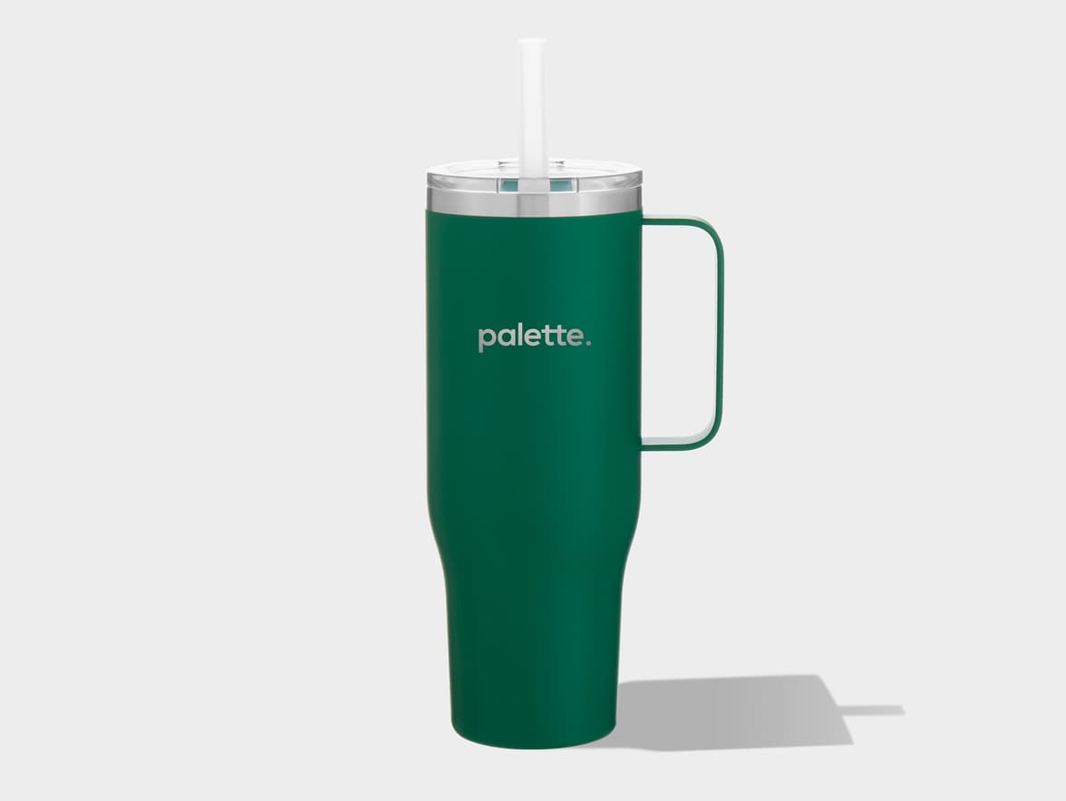 A tall, dark green tumbler with a matching integrated handle on its right side. It has a silver-colored rim and a clear, translucent lid with a white straw inserted in the center. The word 'palette.' is printed in silver, sans-serif text on the front of the tumbler. The item is displayed on a plain light gray background, casting a distinct shadow to its lower right.