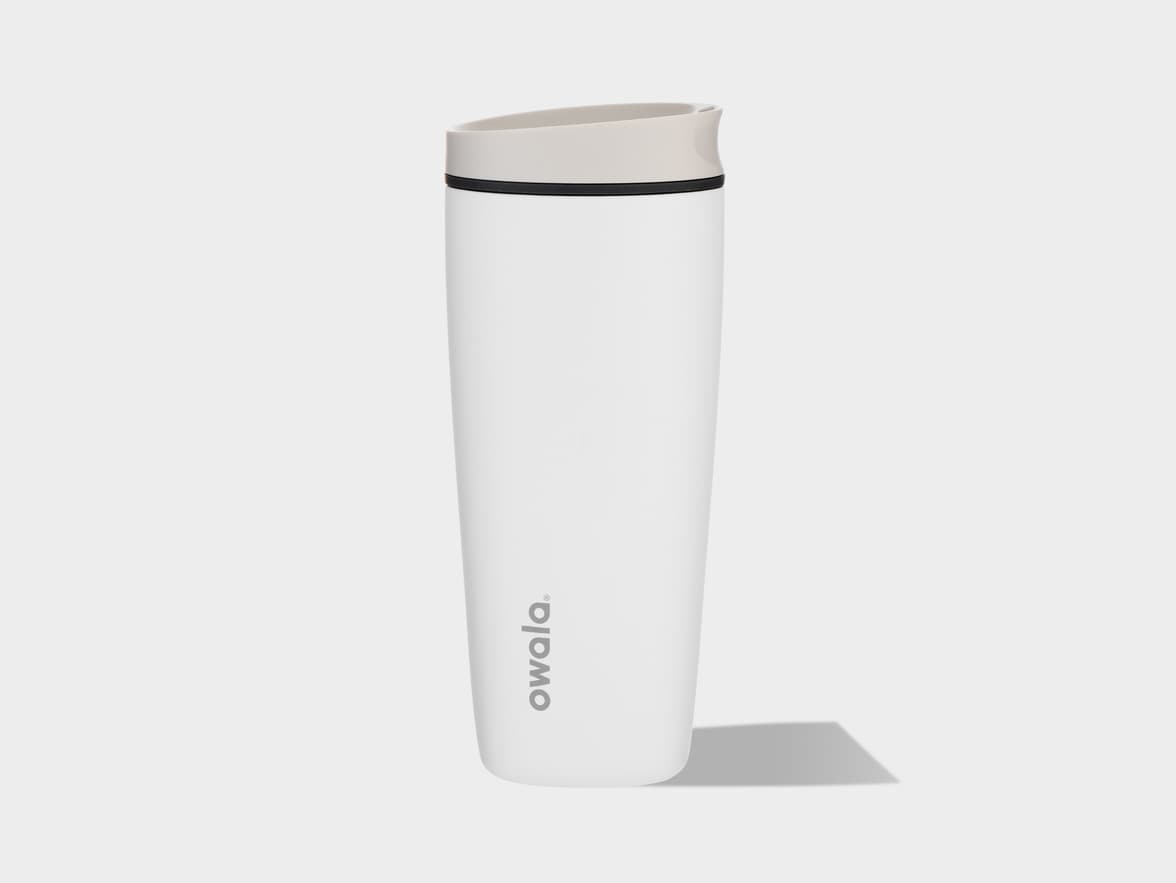A white Owala travel mug with a light grey lid and a black accent ring, standing upright on a light grey surface with a shadow extending to its right. The "owala." logo is oriented vertically on the front of the mug.