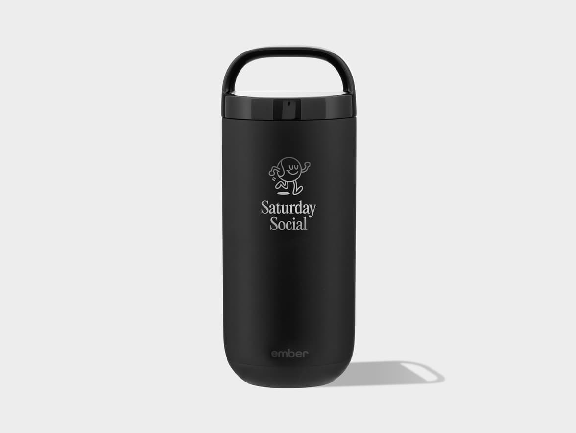 A matte black Ember travel mug with a black handle on its lid. The mug features a light grey outline drawing of a cartoon character with a wavy body and two legs, above the text "Saturday Social" in a similar light grey font. Near the bottom, the "ember" logo is subtly debossed or printed in light grey. The mug stands upright on a light grey background, casting a shadow to its right.