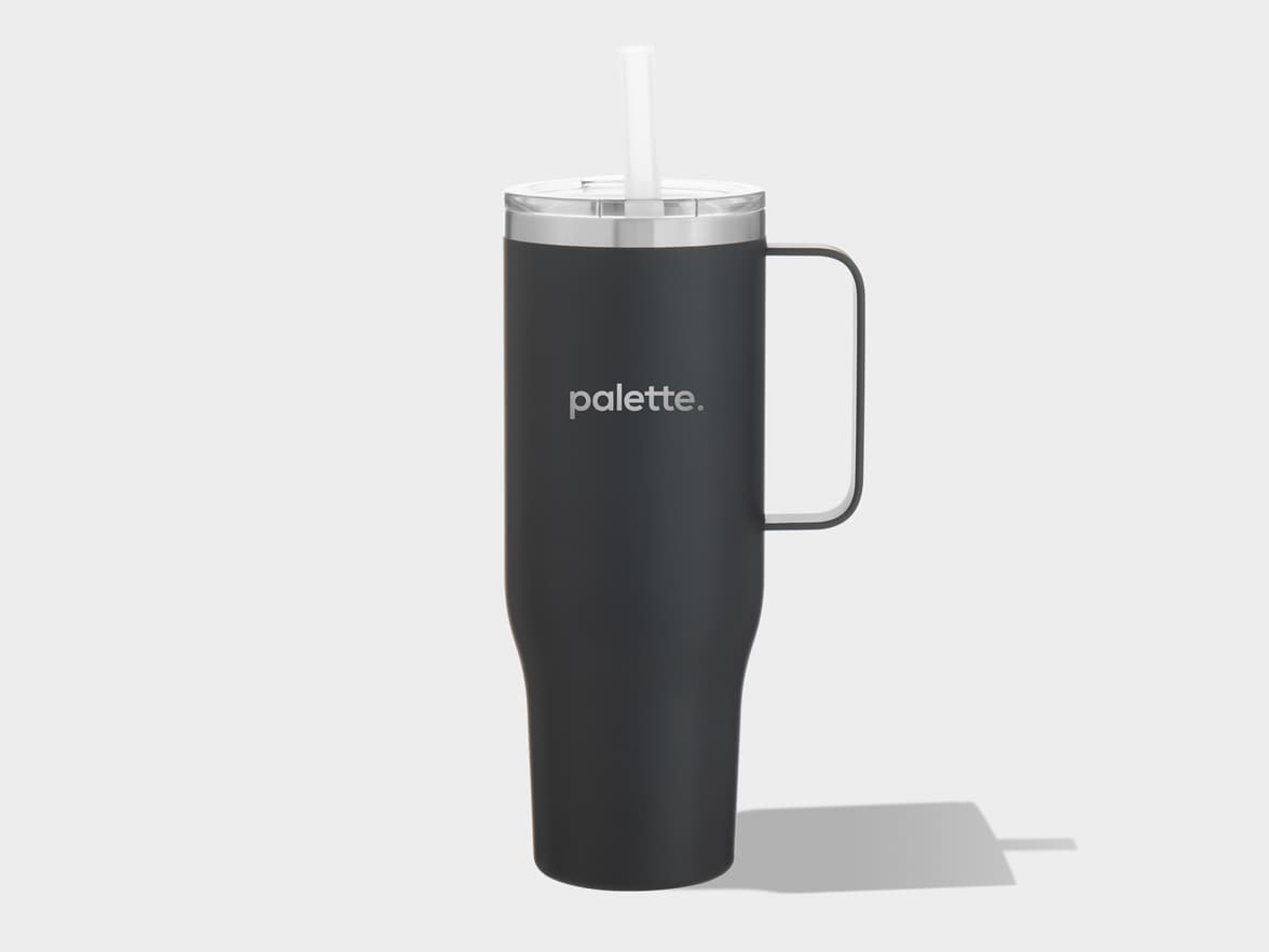 A dark grey travel mug with a handle is positioned centrally on a light grey background. The mug has a silver rim around the top and a clear plastic lid with a transparent straw protruding from it. The word "palette." is printed horizontally in light grey on the front of the mug. A soft, dark grey shadow extends from the base of the mug towards the right side of the frame.