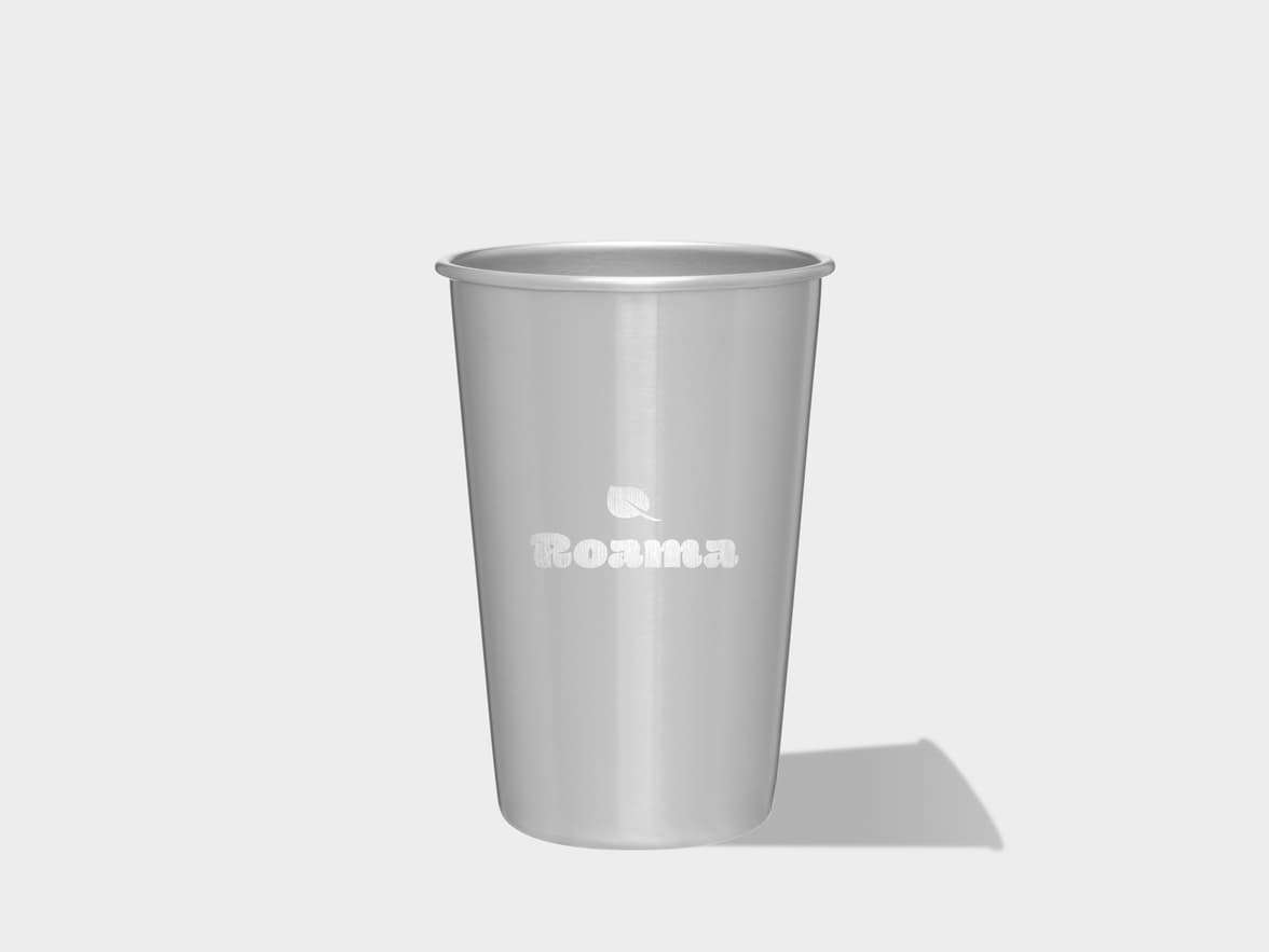 A single, upright silver metal cup with a brushed finish stands on a light gray surface, casting a subtle shadow to its right. The cup features a white "Roama" logo with a small leaf icon above it, centered on its front.																								