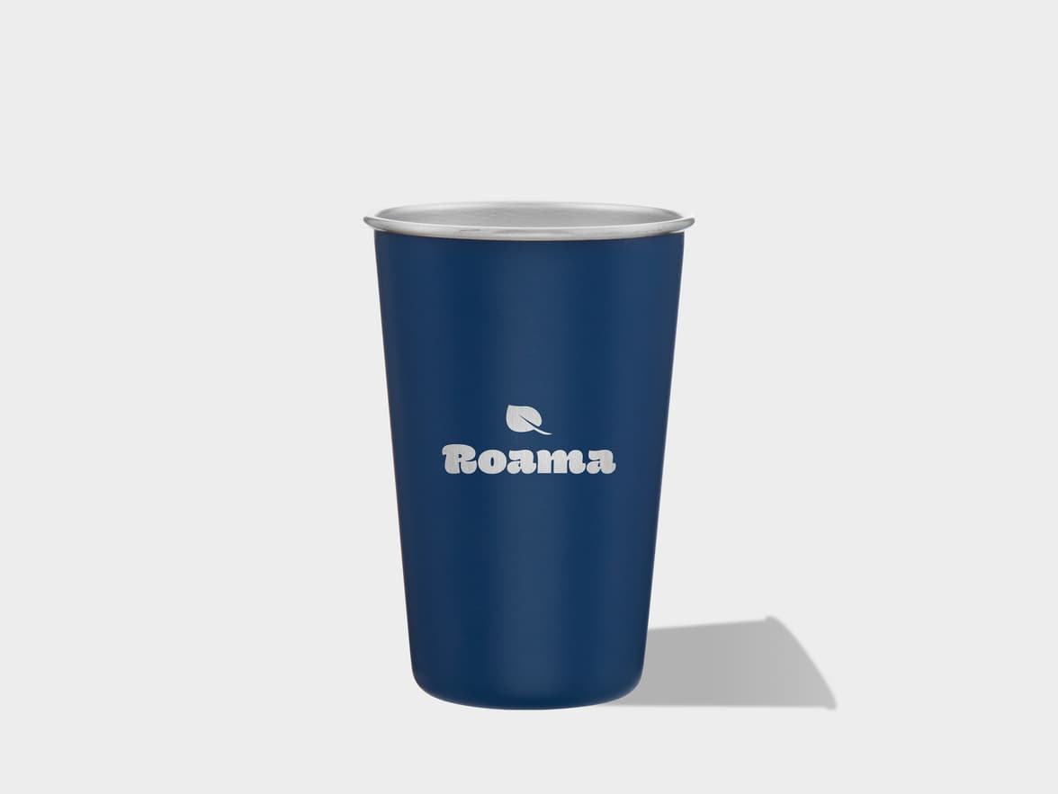 A navy blue travel tumbler with a silver rim stands upright on a light grey surface, casting a shadow to its right. A white logo, featuring a leaf above the word "Roama," is visible on the front of the tumbler.